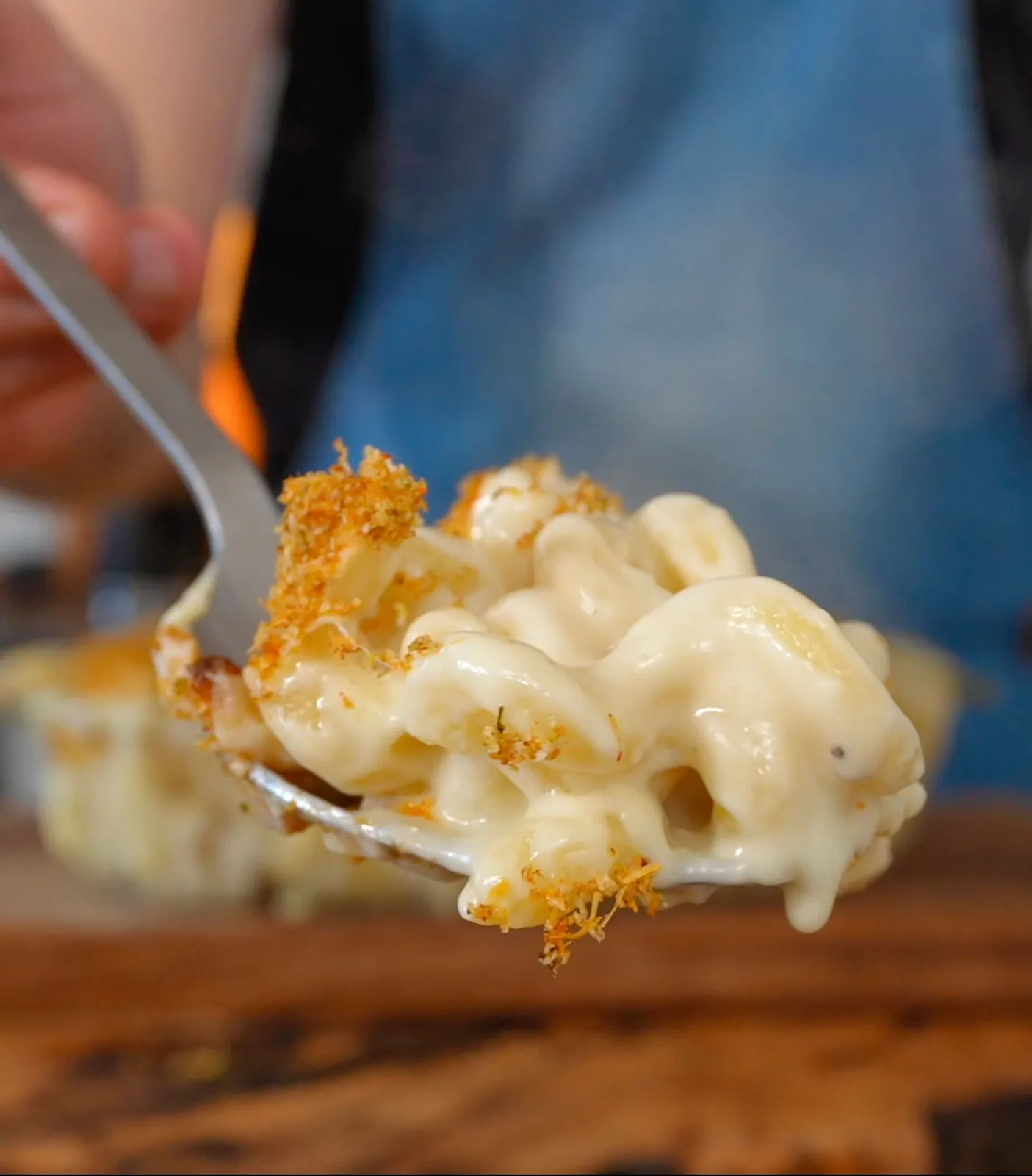 A spoonful of creamy macaroni and cheese with crispy topping, held over a wooden table. Blue denim apron in the blurred background.