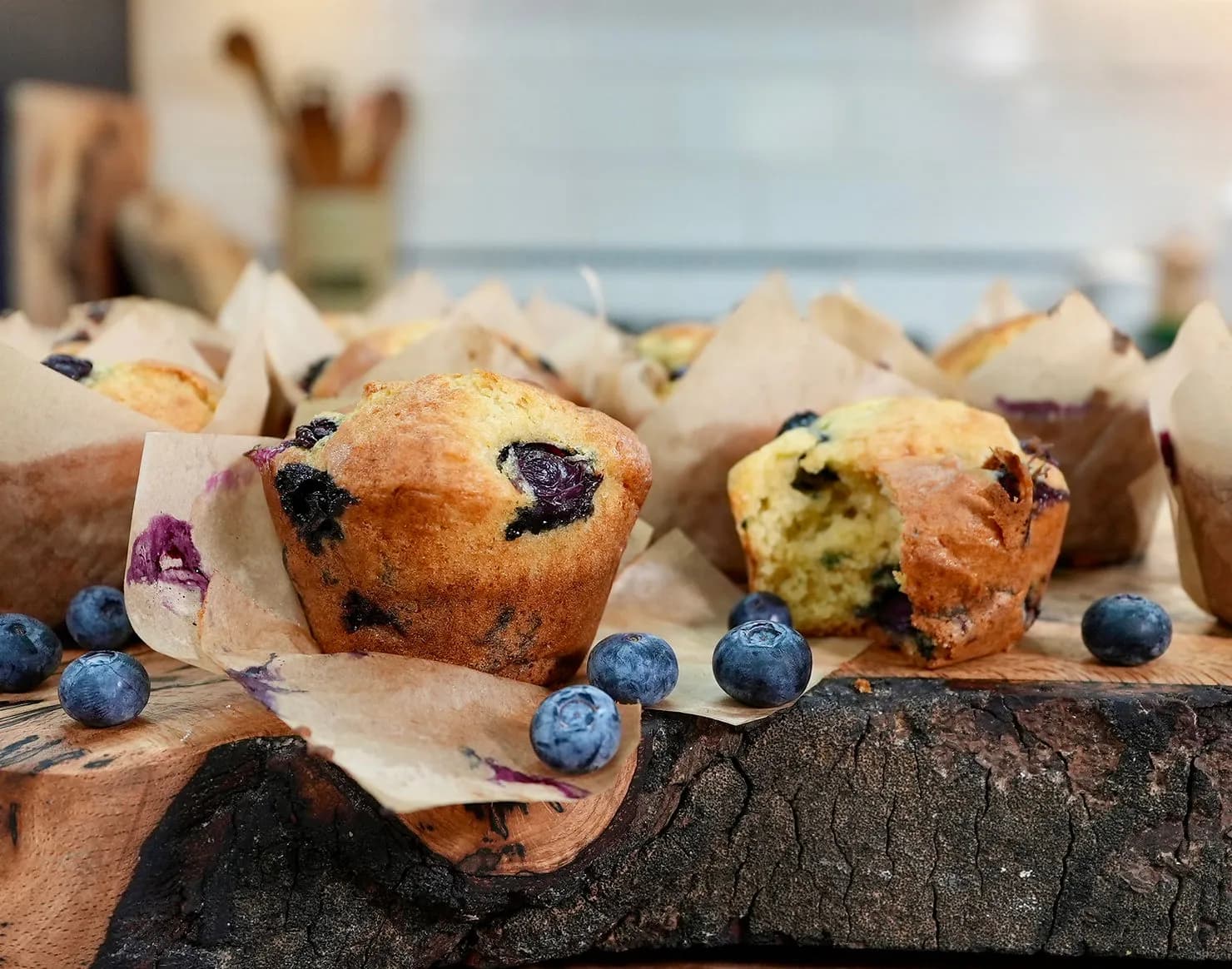 Freshly baked blueberry muffins on a rustic wooden board with scattered blueberries. Warm kitchen setting, creating a cozy, inviting mood.