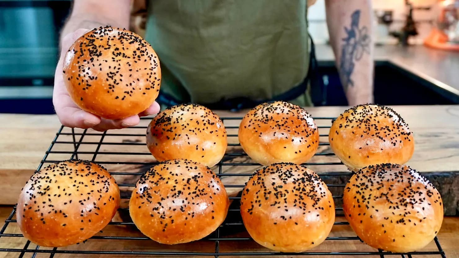 Person holding a burger bun, showcasing seven similar buns on a cooling rack in a kitchen. Bun tops are golden brown with black seeds.