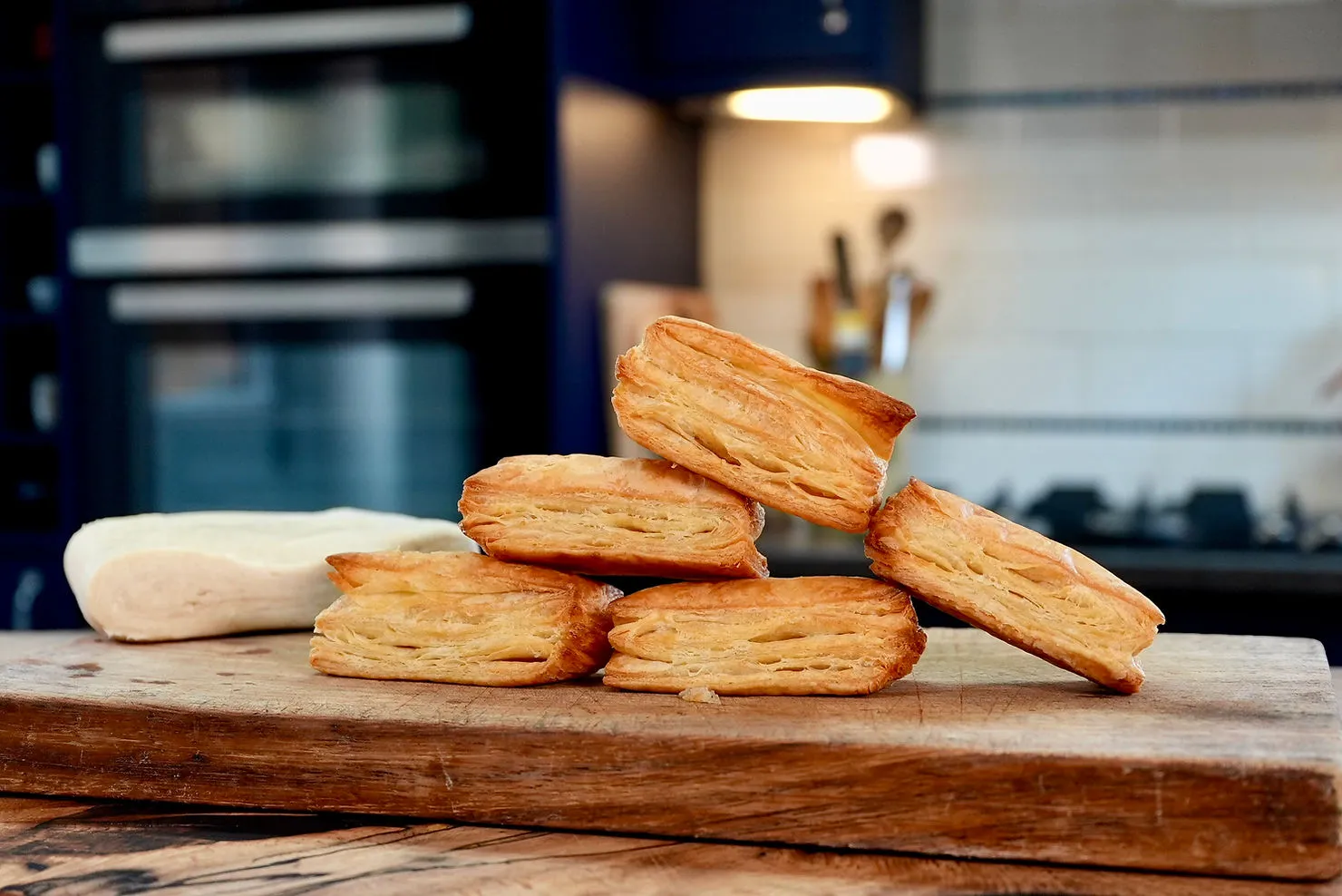 Pastry pieces stacked on a wooden board with dough nearby. Kitchen background with warm lighting and soft focus appliances.