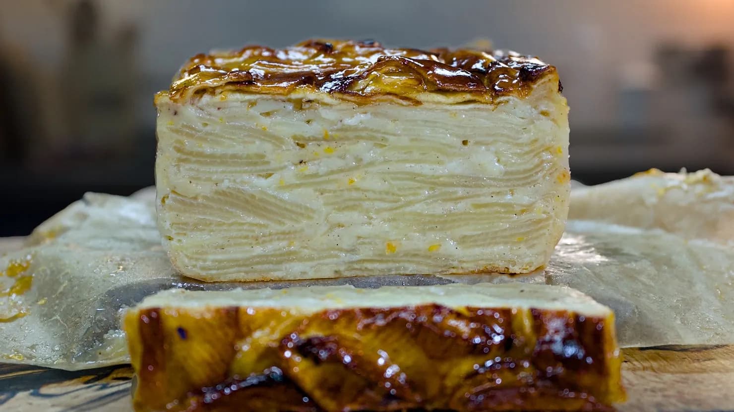 Layered apple cake on parchment paper, with a caramelized top and visible apple layers. Warm kitchen setting, soft lighting.