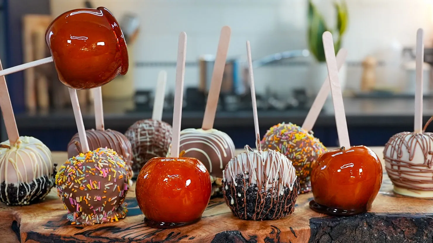 Candy apples with various coatings and colorful sprinkles on a wooden board in a kitchen setting, creating a festive, appetizing display.