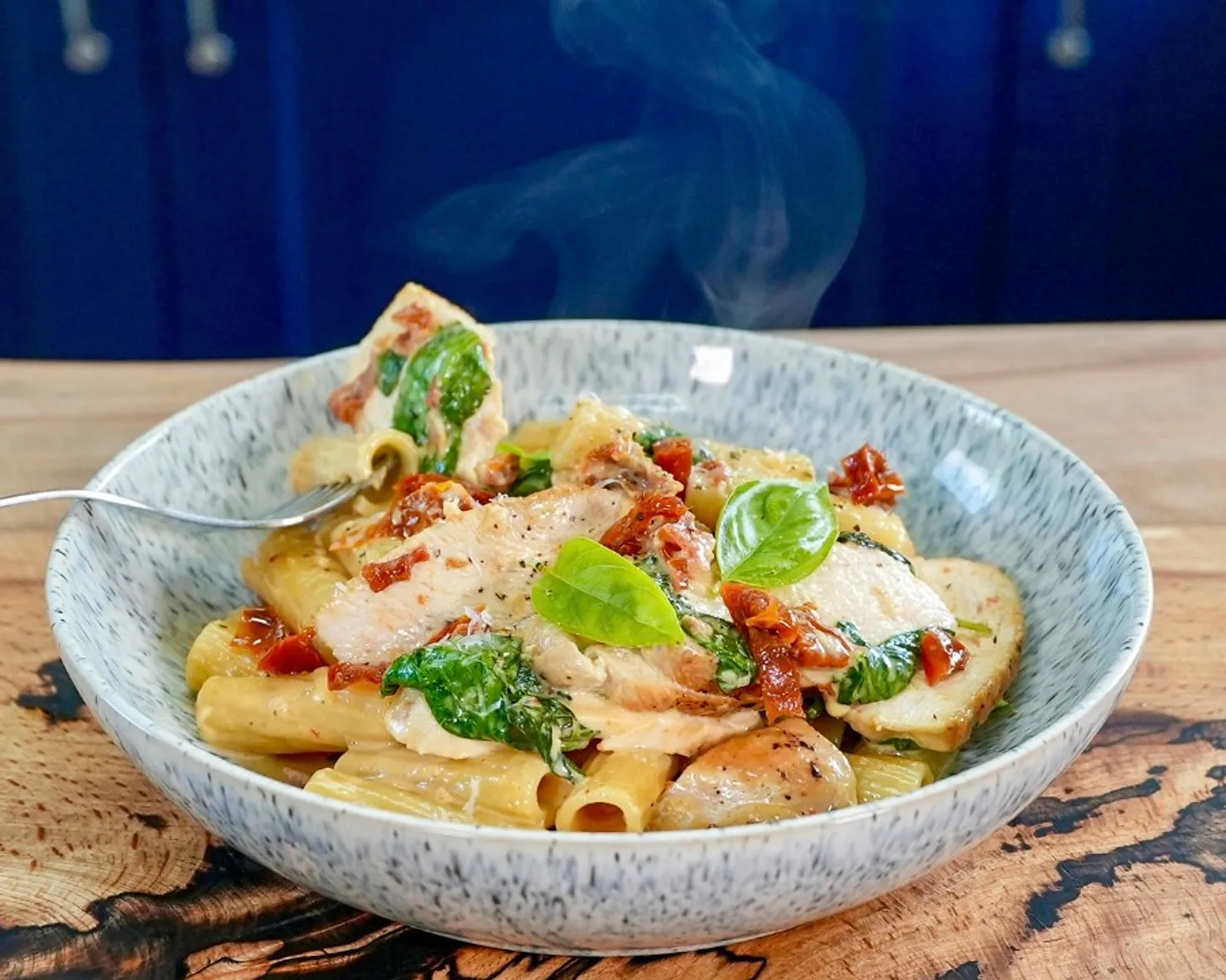 Creamy pasta with chicken, spinach, and sun-dried tomatoes in a blue speckled bowl on a wooden table. Steam rises, creating a warm, inviting mood.