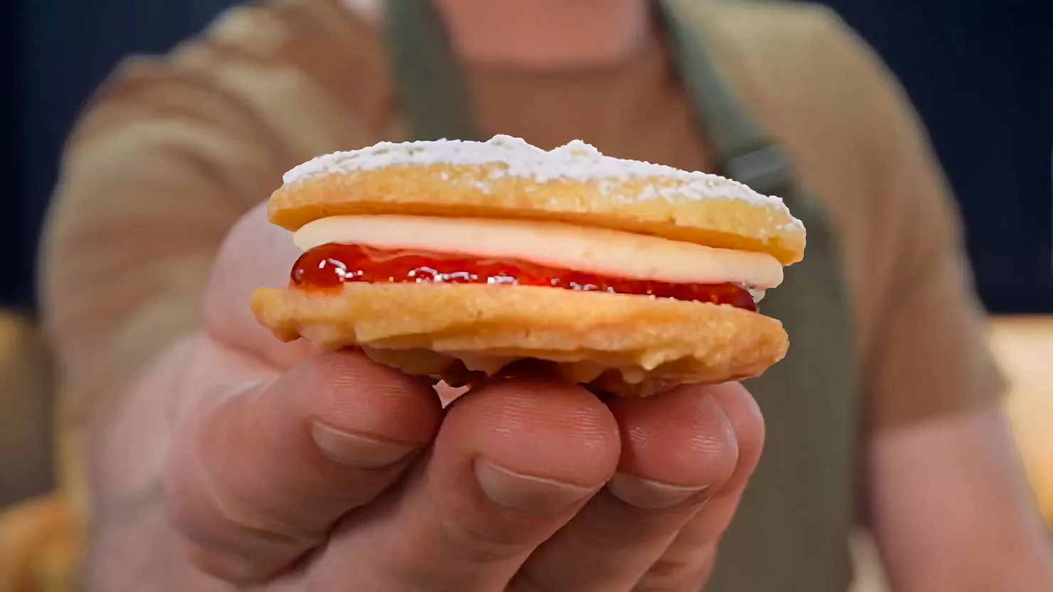 A hand holding a round cookie with powdered sugar, cream, and red jelly filling. Person in brown shirt and apron blurred in background.