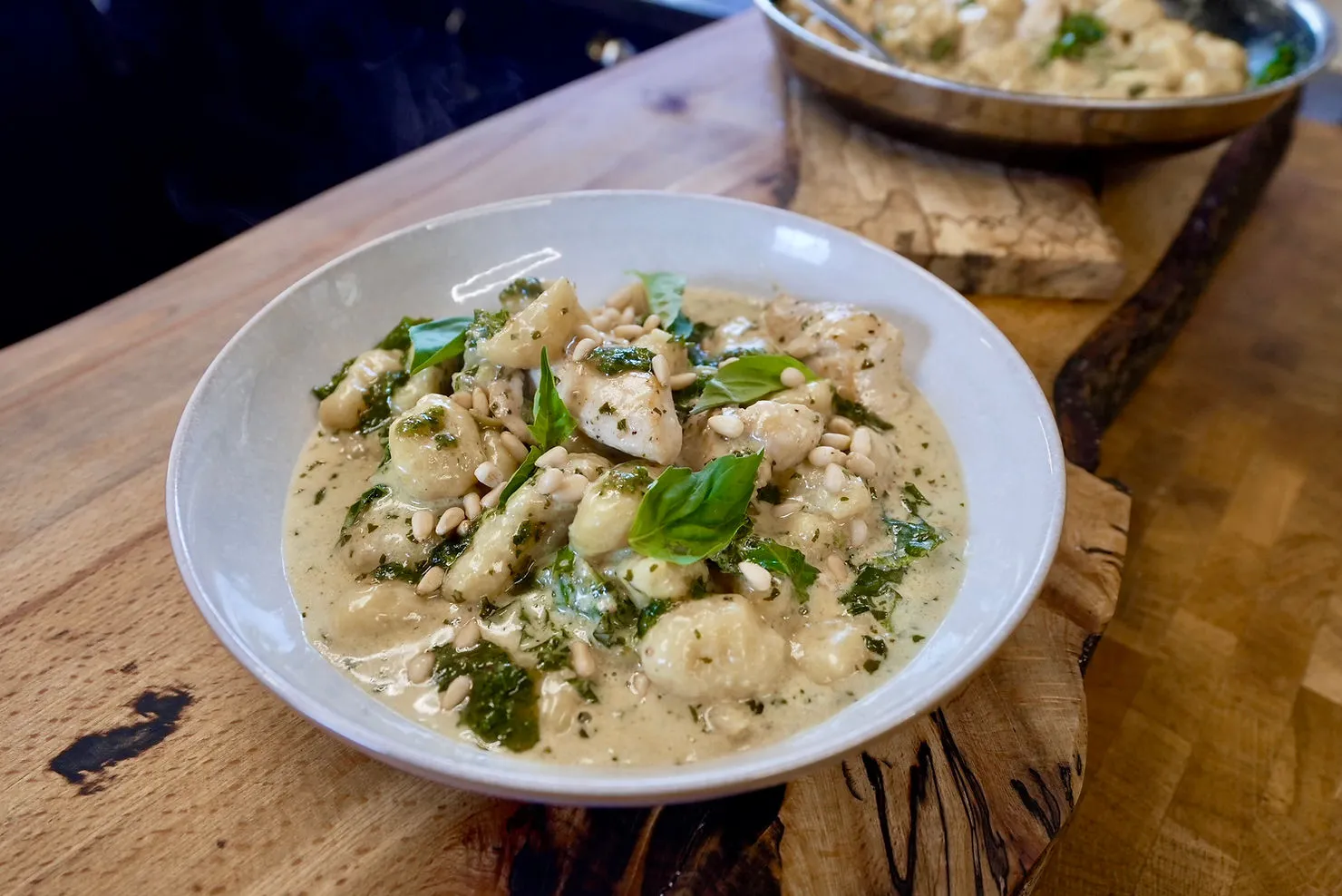 Creamy gnocchi with basil, spinach, and pine nuts in a white bowl on a wooden table. Another dish is partially visible in the background.