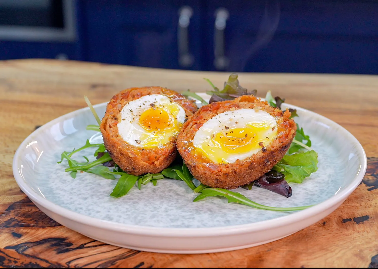 Scotch egg halved on salad greens, served on a white plate. The yolk is vibrant and runny. Wooden table background.
