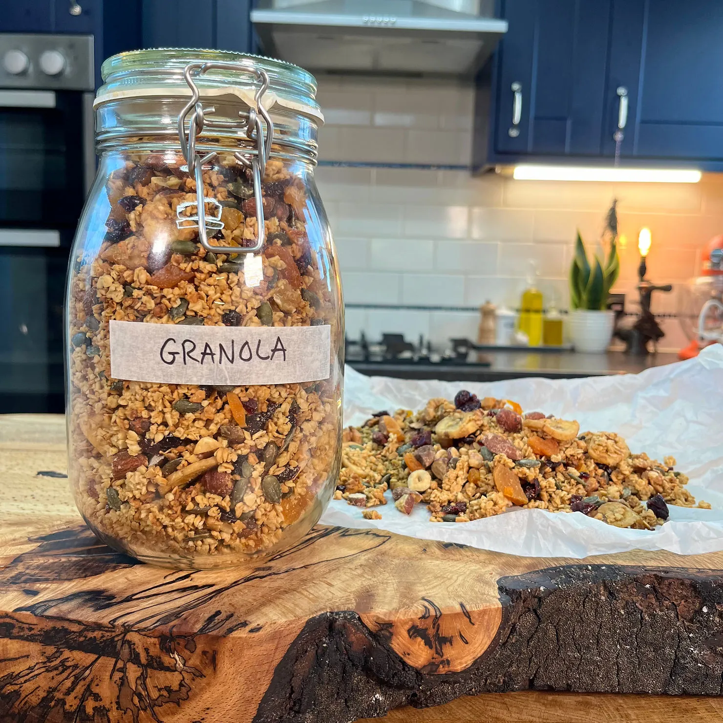 Jar labeled "Granola" on a wooden board, with loose granola beside it. Kitchen setting with blue cabinets and white tiles in background.