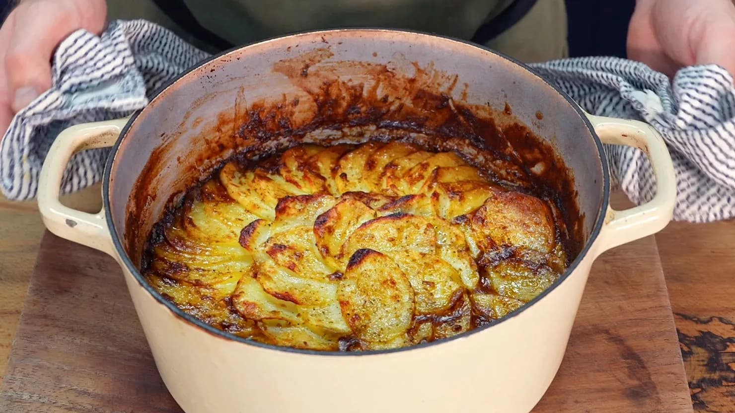 Browned potatoes in a cream-colored pot on a wooden surface. A person in the background holds the pot with a striped cloth. Cozy, rustic feel.