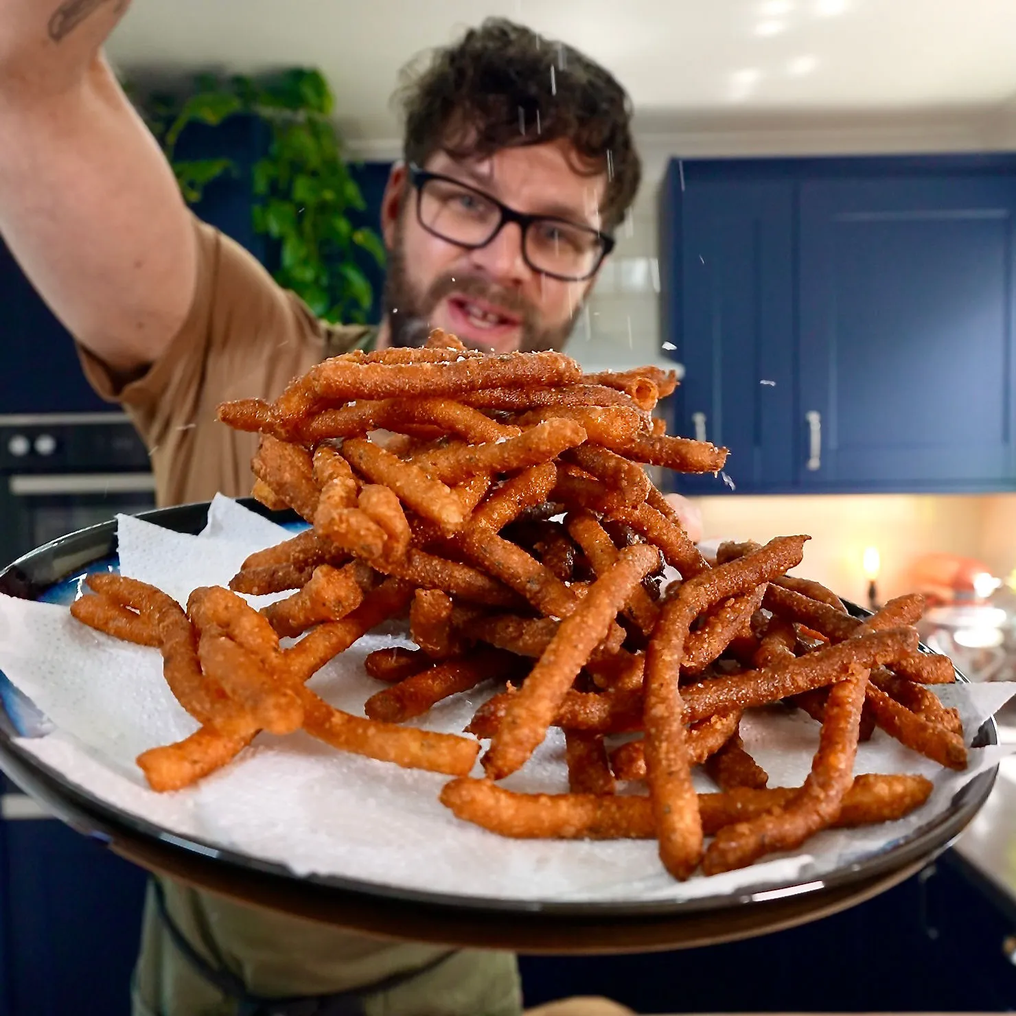 Man sprinkling seasoning on crispy fries in a kitchen with blue cabinets. The fries are served on a paper-lined plate. Energetic vibe.