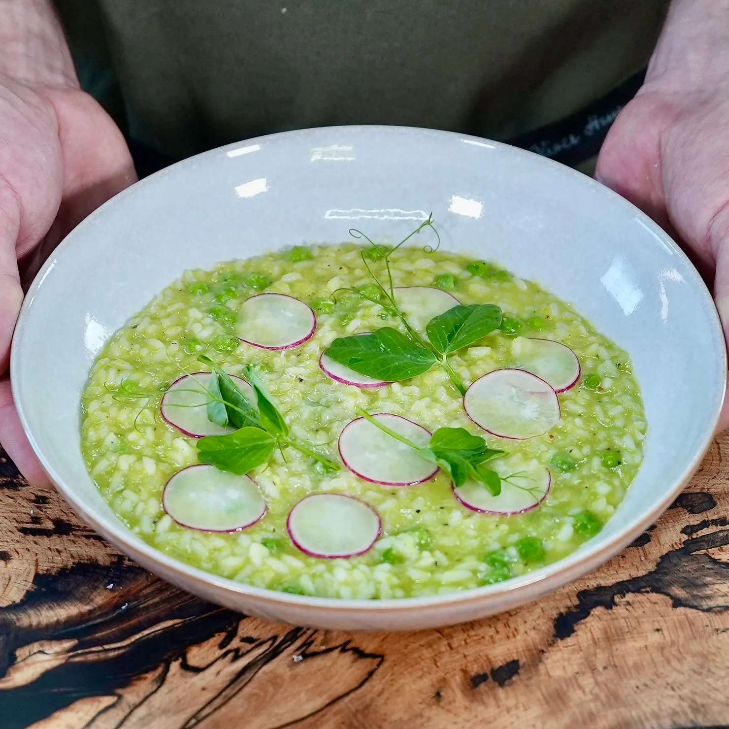 A person holds a bowl of green risotto topped with radish slices and pea shoots, set on a wooden table. The mood is fresh and appetizing.