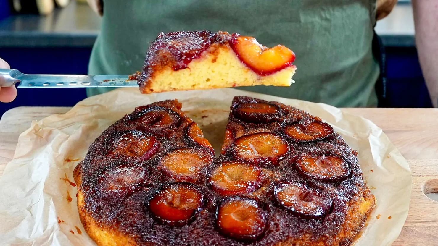 A slice of plum upside-down cake on a spatula, showing moist, caramelized fruit atop a golden-yellow sponge, set on parchment paper.