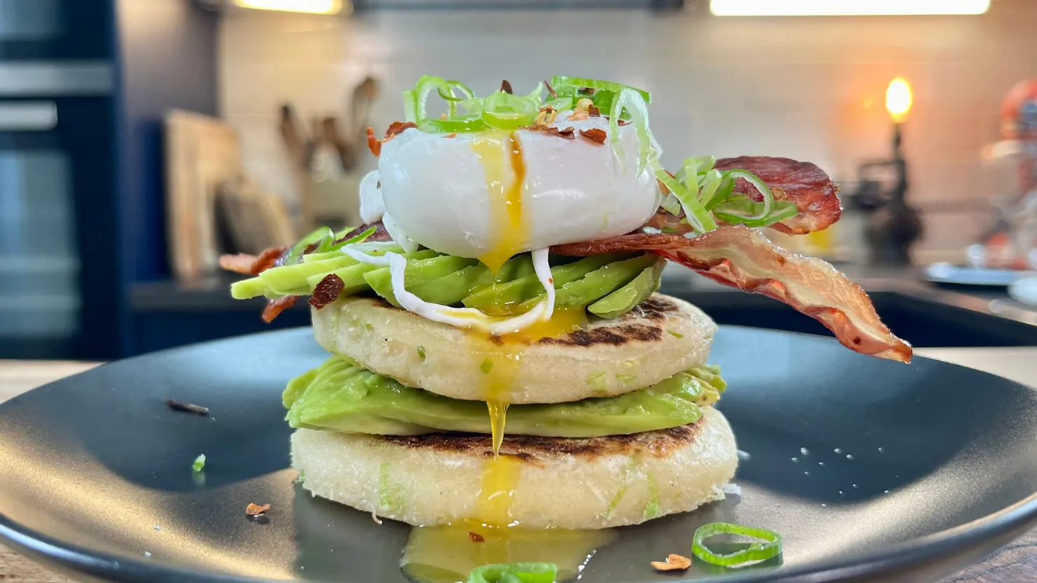 Gourmet dish with poached egg, avocado, and bacon on potato cakes, topped with green onions. Yolk drips on a dark plate in a kitchen setting.
