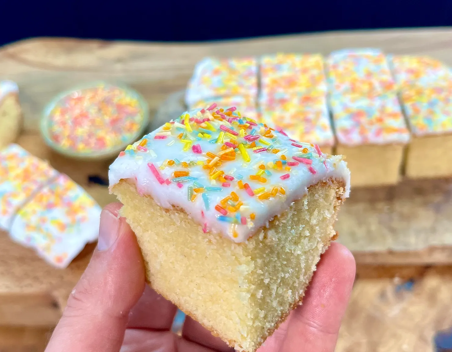A hand holds a frosted vanilla cake slice topped with colorful sprinkles. More slices and a bowl of sprinkles are on a wooden table.