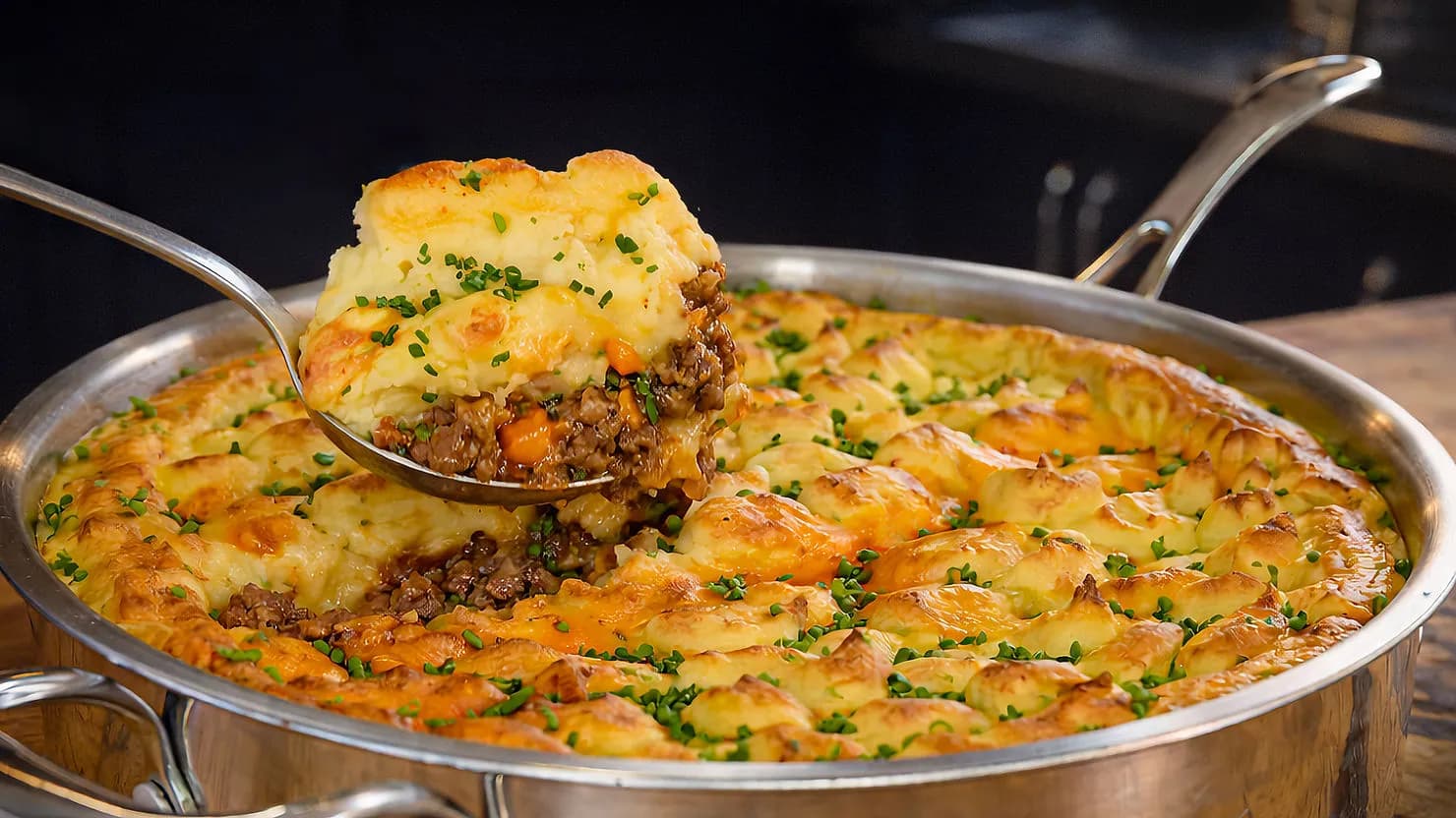 A spoon lifts a portion of shepherd's pie from a silver pan. The pie has golden mashed potatoes and is garnished with chopped chives.