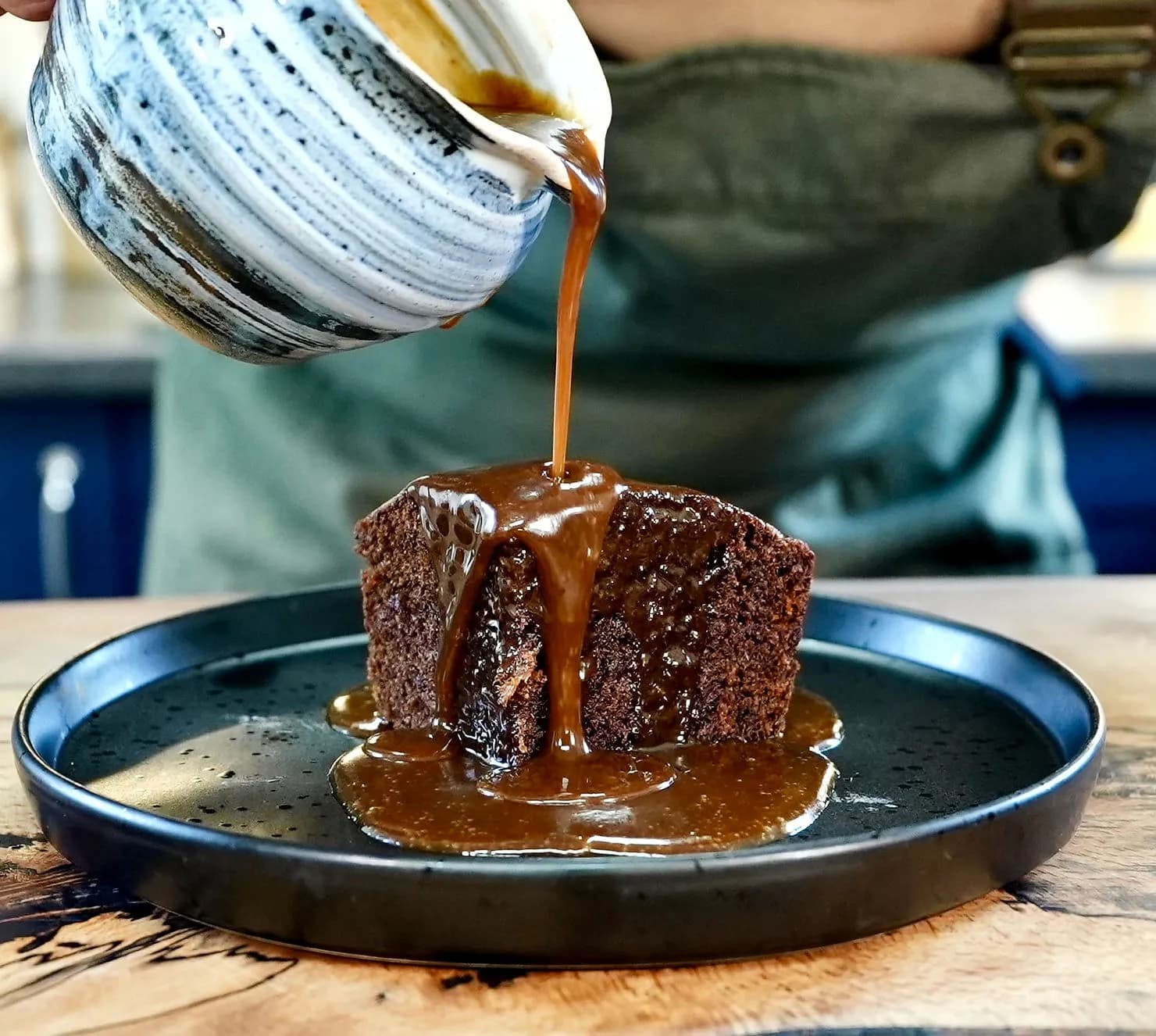 Toffee sauce pours over Sticky Toffee Pudding on a black plate. A person in an apron holds the marbled bowl, creating a cozy kitchen vibe.