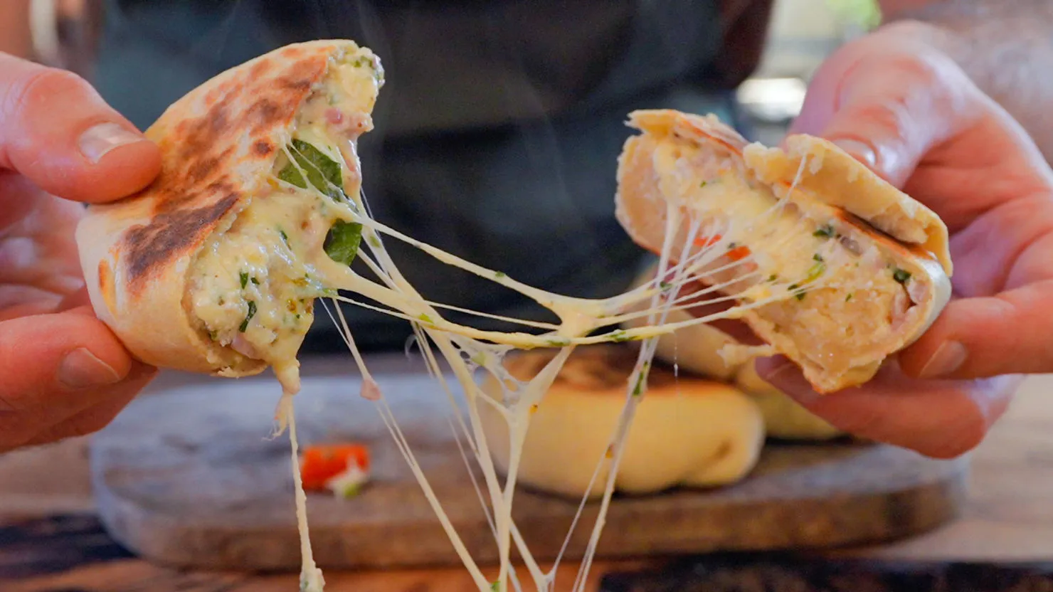 Hands pulling apart gooey, cheesy bread with spinach and ham on a wooden board. Melted cheese stretches between the halves, creating a warm, appetizing mood.