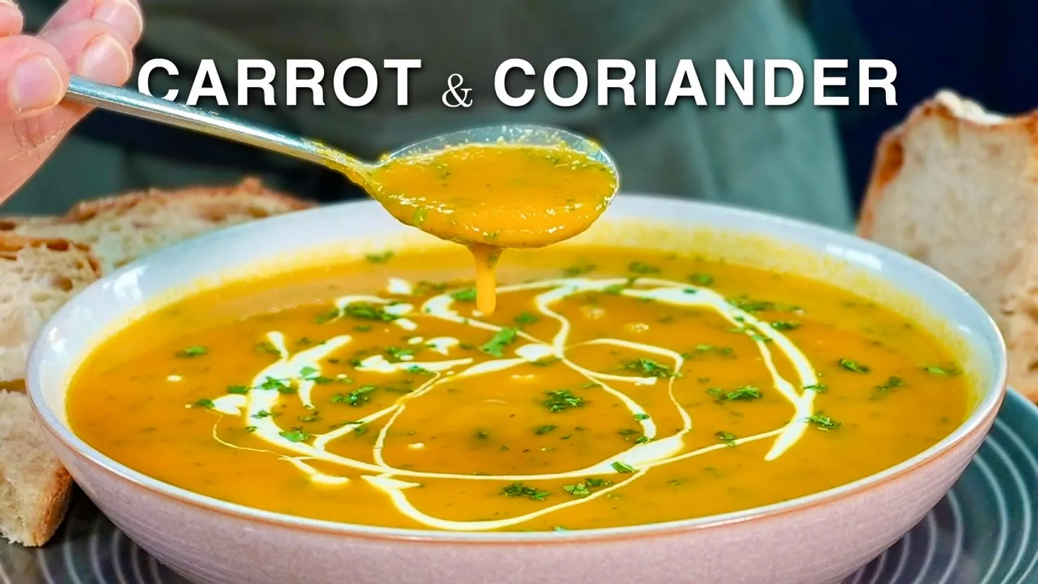 Bowl of carrot and coriander soup with cream swirls and herbs. A spoon lifts soup above the bowl. Bread in the background. Text: Carrot & Coriander.