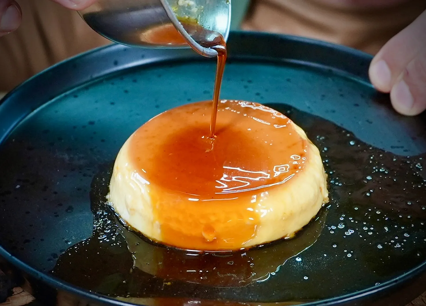 Caramel sauce is being poured over a round flan on a dark speckled plate. The scene conveys a tempting dessert preparation.
